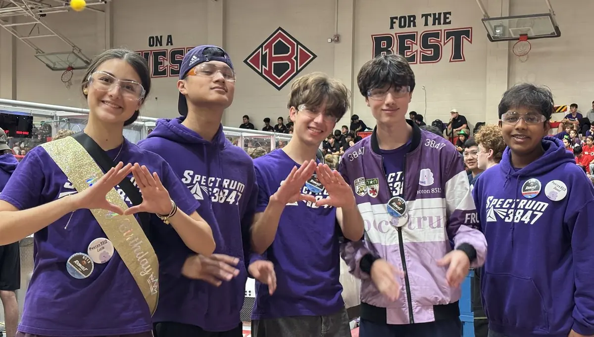 Five Spectrum students in purple team shirts at a competition, making the team hand sign